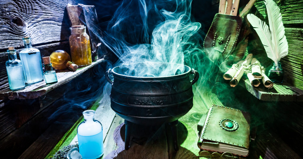 A cauldron filled with pale blue smoke sits amidst other accoutrements of witchcraft, shrouded in blue and green light.