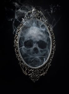 A mirror in an ornate frame against a black background, within the mirror a skull made of smoke lingers.