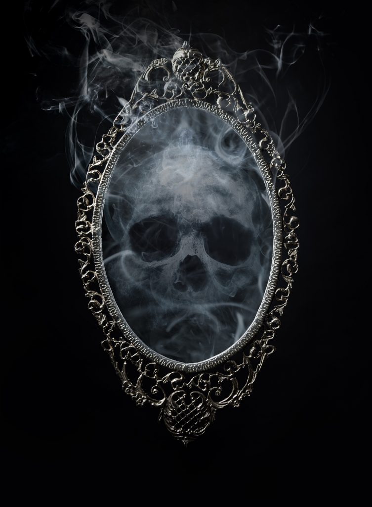 A mirror in an ornate frame against a black background, within the mirror a skull made of smoke lingers.