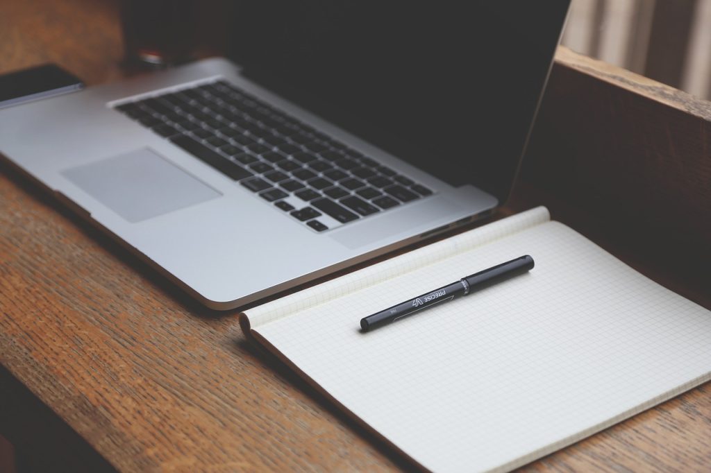 A laptop sits on a desk, an empty notepad lying open next to it.